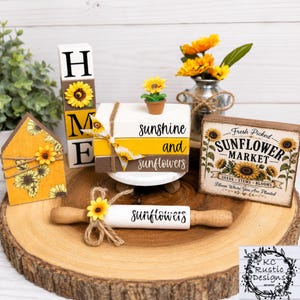 May include: A sunflower-themed decor set featuring a wooden house sign, a vertical HOME block, a stack of three books with twine and a sunflower, a Fresh Picked Sunflower Market sign, and a white rolling pin with the word sunflowers printed on it.