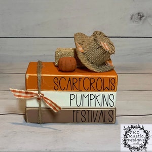 Peut inclure: Une pile de trois blocs en bois peints en orange, blanc et marron. Les blocs sont décorés d'une inscription noire qui dit "Scarecrows", "Pumpkins", et "Festivals". Un petit chapeau de paille de scarecrow brun se trouve sur le dessus des blocs.