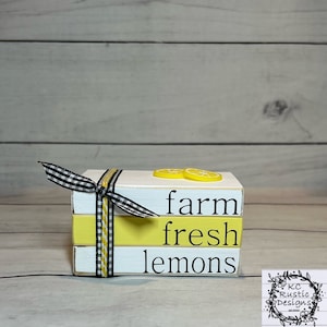 May include: A stack of three white wooden blocks with black lettering that reads "farm fresh lemons". The blocks are tied together with a black and white checkered ribbon and a yellow ribbon. Two yellow lemon slices are on top of the blocks.