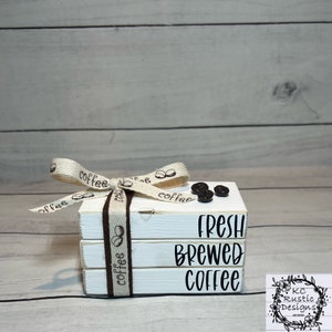 May include: A stack of three white wooden blocks with the words "Fresh Brewed Coffee" written in black. The blocks are tied together with a brown ribbon with the word "coffee" printed on it. There are three coffee beans on top of the blocks.