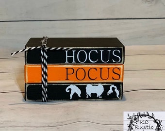 May include: A three-tiered wooden block set with the words "Hocus Pocus" painted in white on a black background. The bottom block has three white silhouettes of witches. The blocks are tied together with black and white twine.