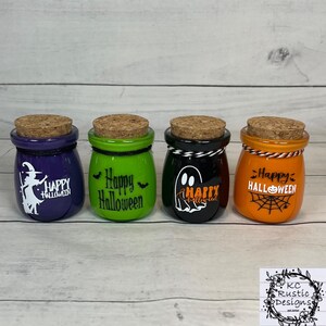 Könnte beinhalten: Vier kleine, farbige Glasbehälter mit Korken, die für Halloween dekoriert sind. Die Gläser sind lila, grün, schwarz und orange, mit weißem und orangefarbenem Text und Grafiken. Jedes Glas hat ein anderes Design, darunter eine Hexe, Fledermäuse, ein Geist und ein Spinnennetz.