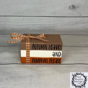 Könnte beinhalten: Dekoratives Holzblock-Set mit den Worten "AUTUMN LEAVES AND PUMPKINS PLEASE" in schwarzer Schrift. Die Blöcke sind braun, weiß und orange, gebunden mit einem braun-weiß karierten Band.
