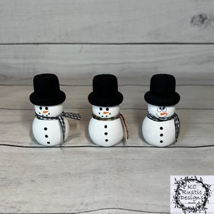 May include: Three white painted snowman figurines with black top hats and black and white scarves. The snowmen have black eyes and orange carrot noses.