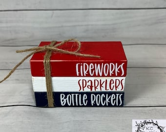 May include: A stack of three wooden blocks painted red, white, and blue, tied with twine. The top block reads "FIREWORKS", the middle "SPARKLERS", and the bottom "BOTTLE ROCKETS" in white script. A rustic design.