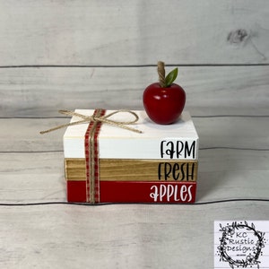 May include: A stack of three wooden blocks with the words "Farm Fresh Apples" painted on them. The top block is white, the middle block is brown, and the bottom block is red. A red apple with a green stem and leaf sits on top of the stack.