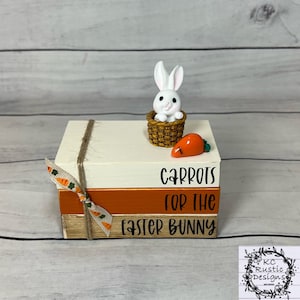 Op de afbeelding: Een decoratieve paasstapel van drie houten blokken. Het bovenste blok is wit, het middelste is oranje en de onderste is houtkleurig. Een wit konijn in een mand en een oranje wortel staan bovenop de blokken. De tekst luidt "Carrots for the Easter Bunny."