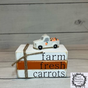 May include: A white wooden tiered sign with the words "farm fresh carrots" stacked on top of each other. A small white truck with orange carrots in the bed sits on top of the sign.