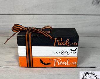 May include: A set of three wooden blocks stacked on top of each other. The top block is black and says "Trick" in orange lettering. The middle block is white and says "or" in black lettering. The bottom block is orange and says "Treat" in black lettering. Each block has a black bat silhouette.