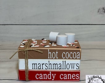Könnte beinhalten: Stapelbare Holzblöcke mit den Worten "hot cocoa", "marshmallows" und "candy canes" darauf bemalt. Der oberste Block hat weiße Marshmallows und rote und weiße Streusel.