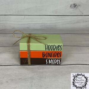 May include: A stack of three wooden blocks bound with twine. The top block is light green, the middle is orange, and the bottom is brown. The words "Hoodies", "Bonfires", and "S'mores" are written on the blocks in black.