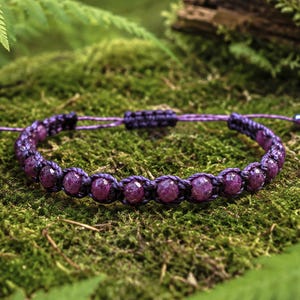 Könnte beinhalten: Ein violettes Kordelarmband mit einer Reihe facettierter, tief magentafarbener Perlen. Das Armband ist in Makramee-Knotentechnik gefertigt und verfügt über einen verstellbaren Schiebeknotenverschluss mit zwei kleinen Perlen an den Enden.