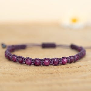 Könnte beinhalten: Nahaufnahme eines handgefertigten Armbands mit kleinen, runden, tiefroten Edelsteinen. Die Edelsteine sind mit einer dunkelvioletten Kordel aufgereiht, wodurch ein Makramee-Design entsteht. Das Armband liegt auf einer hellbraunen Holzoberfläche.