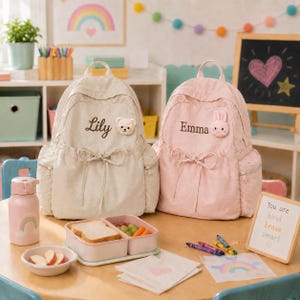 May include: Two backpacks in cream and light pink, each featuring a drawstring front, side pockets, and a plush animal patch. The cream bag is embroidered with the name Lily and a bear, while the pink bag displays the name Emma and a rabbit.