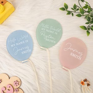 Puede incluir: Tres placas con nombres en forma de globo de colores pastel. Cada placa tiene el texto "Hello World, My Name Is" seguido de un nombre y un apellido. Los globos son azules, verdes y rosas, con texto y cuerda blancos.