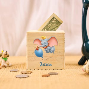 Puede incluir: Huchita de madera con una imagen de dibujos animados de Dumbo el elefante sosteniendo un globo azul. El nombre "Karen" está impreso en la parte delantera de la huchita.