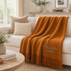 Puede incluir: Una manta texturizada de color naranja quemado con flecos, que presenta el nombre Charlotte bordado en letra cursiva azul claro sobre un peque&ntilde;o dise&ntilde;o de hojas. La manta est&aacute; colocada sobre un sof&aacute; de color claro con dos cojines decorativos.