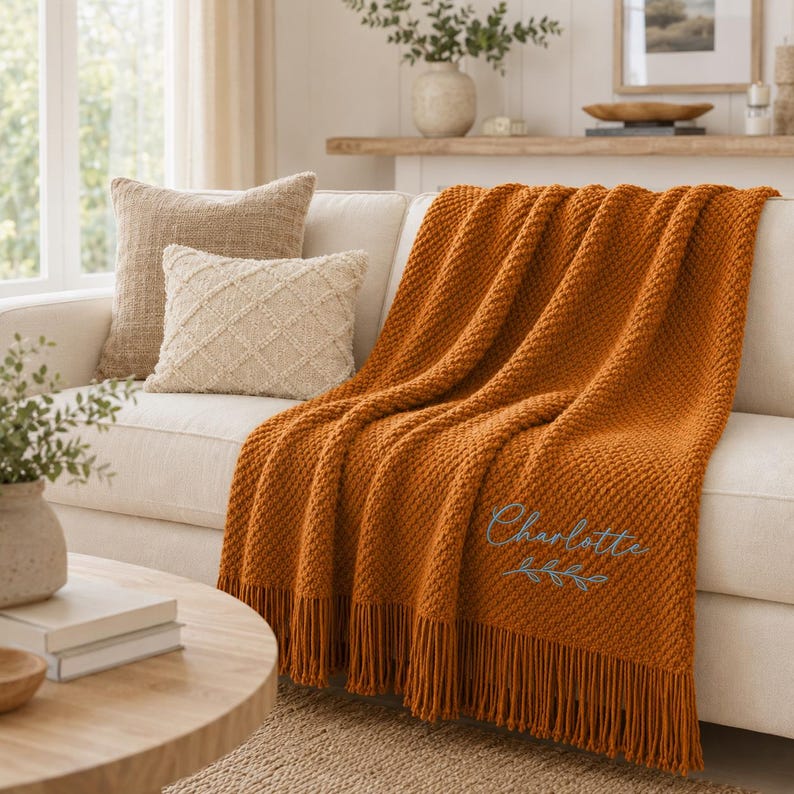 Puede incluir: Una manta texturizada de color naranja quemado con flecos, que presenta el nombre Charlotte bordado en letra cursiva azul claro sobre un peque&ntilde;o dise&ntilde;o de hojas. La manta est&aacute; colocada sobre un sof&aacute; de color claro con dos cojines decorativos.