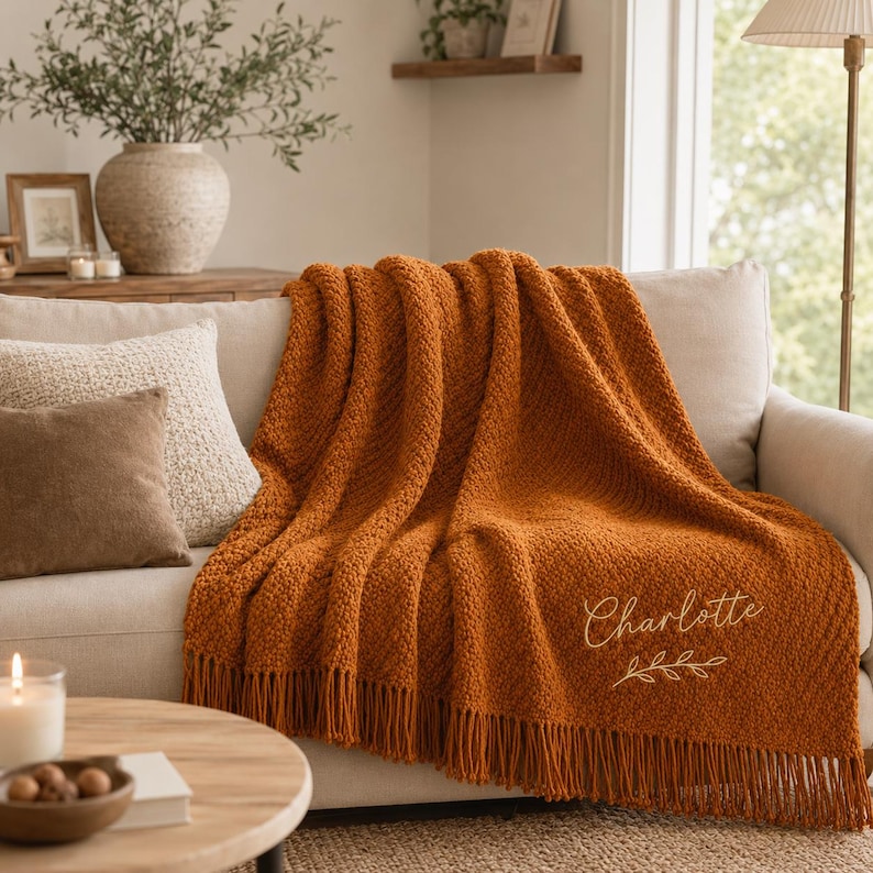 Puede incluir: Una manta tejida de color naranja quemado con flecos, que presenta el nombre Charlotte bordado en letra cursiva sobre un peque&ntilde;o dise&ntilde;o de rama de hojas, colocada sobre un sof&aacute; de color claro con cojines texturizados.