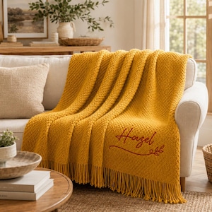 Puede incluir: Una manta de punto grueso color amarillo mostaza con un patr&oacute;n de textura y flecos largos en el borde inferior. El nombre Hazel est&aacute; bordado en una letra cursiva de color rojo oscuro con un adorno decorativo en la esquina inferior derecha.