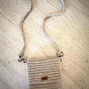 May include: A beige crochet crossbody bag with a matching strap. The bag has a rectangular shape with a flap closure and a small brown leather tag. The strap is attached with metal clasps.
