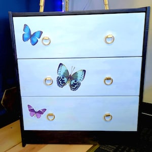 May include: A three-drawer chest of drawers with a light blue painted finish. The drawers feature butterfly decals in blue and purple, and gold-colored ring pulls. The chest has a black frame and base.