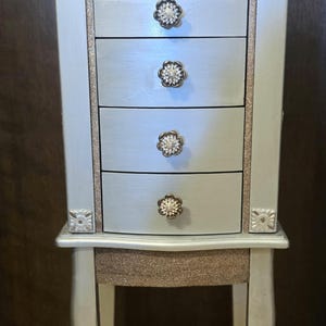 May include: A light blue jewelry armoire with five drawers and silver glitter accents. The armoire has flower-shaped drawer pulls and decorative corner details. The legs are curved and painted silver.