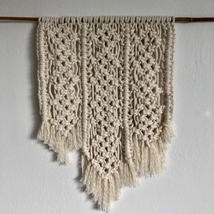 May include: A beige macrame wall hanging with a geometric pattern and fringe. The hanging is attached to a wooden dowel.