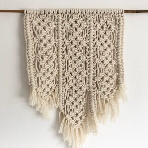 May include: A beige macrame wall hanging with a geometric pattern and fringe details.
