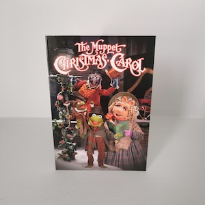 May include: A promotional image for "The Muppet Christmas Carol" shows the Muppets in festive attire. Gonzo in a red coat and top hat, Kermit and Fozzie in winter coats, and Miss Piggy in a bonnet and plaid dress. The title is in a decorative font.