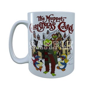 May include: White ceramic mug featuring the title "The Muppet Christmas Carol" in red and brown script. The mug showcases a colorful illustration of Kermit the Frog and other Muppet characters in a winter scene.
