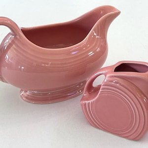 May include: A set of two pink ceramic serving dishes. The larger dish is a gravy boat with a handle and a wide, rounded base. The smaller dish is a pitcher with a handle and a rounded body.