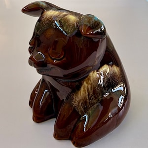 May include: A brown ceramic pig figurine with a glossy finish. The pig is sitting with its front paws forward and has a detailed snout and ears. The glaze has streaks of lighter brown and cream colors.