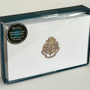 May include: A boxed Harry Potter note card set. The box is dark teal with a clear plastic top. The note cards are white with a gold and purple crest. A circular sticker reads "This Note Card Set Includes: 10 blank note cards, 10 envelopes."