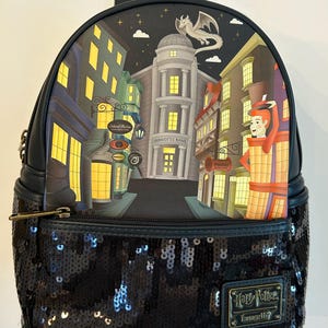 May include: Black sequined backpack featuring a detailed illustration of a city street scene with buildings, a dragon, and the words "Gringotts Bank." The backpack has a top handle, a zippered front pocket, and a Harry Potter Loungefly logo.