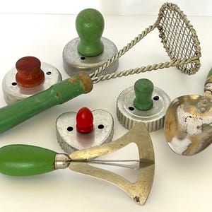May include: A collection of vintage kitchen tools, including cookie cutters, a potato masher, and a scoop. The tools feature wooden handles in green and red, with metal components. The items are arranged on a white surface.