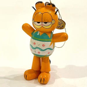 May include: A wooden Garfield figurine with outstretched arms. The orange cat has a white and teal egg-shaped body with pink polka dots. The figurine has black whiskers and a gold string for hanging.