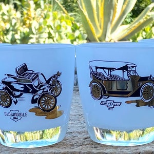 May include: Set of four white glass tumblers with gold trim. Each tumbler features a vintage car illustration with the car's make and year. The cars are a 1902 Rambler, a 1904 Oldsmobile, a 1912 Chevrolet, and a 1903 Ford.