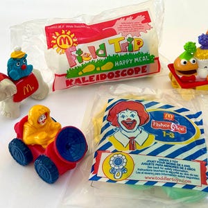 May include: A collection of McDonald's Happy Meal toys, including an elephant, a car with a figure, and a train with characters. The image also shows a sealed bag with a kaleidoscope and a Fisher-Price toy package.