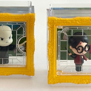 May include: Two small, clear plastic display cases with yellow frames. One contains a bald figure in black robes, the other a figure with glasses and a red and black outfit. Both figures are cartoon-like.