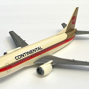 May include: A scale model airplane featuring a cream-colored fuselage with the word CONTINENTAL printed in black. The aircraft has grey wings and engines, with a red and gold stripe running along the side and a red circular logo on the tail fin.