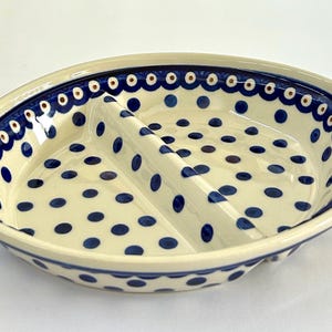 May include: Oval ceramic serving dish with two compartments. The dish is cream-colored with a blue dotted pattern and a scalloped blue border. The dish is designed for serving appetizers or snacks.