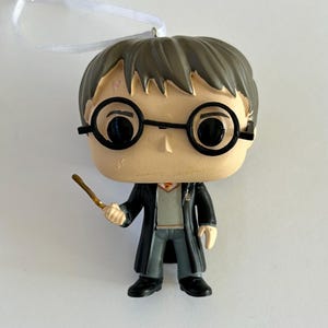 May include: A Harry Potter-themed figurine with a wand, round glasses, and a scar on its forehead. The figure is wearing a black robe, gray pants, and a gray sweater. A white ribbon is attached to the top of the figurine.