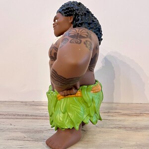 Puede incluir: Una figura de un personaje musculoso con piel oscura, cabello negro rizado y tatuajes tribales. Lleva una falda verde de hojas y se para con las manos en las caderas. La figura es un personaje de dibujos animados.