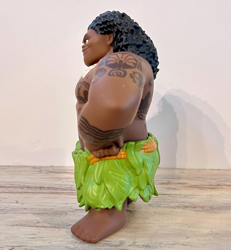 Puede incluir: Una figura de un personaje musculoso con piel oscura, cabello negro rizado y tatuajes tribales. Lleva una falda verde de hojas y se para con las manos en las caderas. La figura es un personaje de dibujos animados.