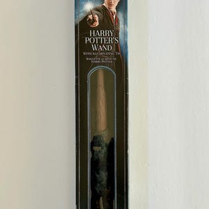 May include: A Harry Potter wand in a display box. The box features an image of Harry Potter and the text "Harry Potter's Wand with Illuminating Tip". The wand is brown with a dark handle. The box has a blue and gold design.