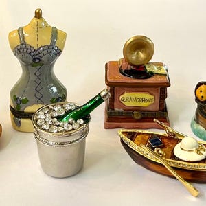May include: A collection of decorative trinket boxes in various shapes, including a teacup, a corset, a champagne bucket, a gramophone, a boat, and a Noah's Ark. The boxes are painted in bright colors and feature intricate details.