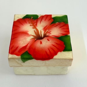 May include: A small, square box with a cream-colored base and lid. The lid is decorated with a vibrant red and white hibiscus flower design, set against green leaves. The box is likely used for jewelry or small keepsakes.