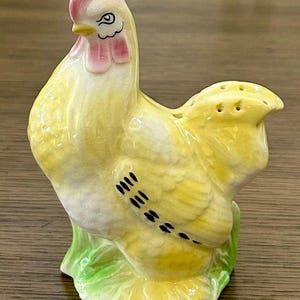 May include: A ceramic rooster-shaped salt shaker. The rooster is primarily yellow with a pink comb and wattle. Black details define the eyes and feather markings. The base is green, and the shaker has three holes on top.