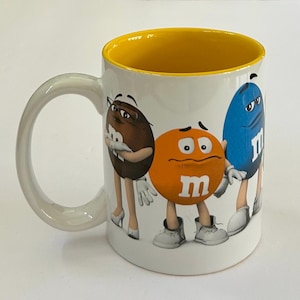 May include: A white ceramic mug with a bright yellow interior and a large handle. The exterior features a graphic print of three M&M characters: a brown one wearing glasses, an orange one with a worried expression, and a blue one standing tall.